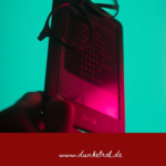 E-Reader mit aufgelegter Brille in rötlichem Licht vor türkisem Hintergrund, darunter der Schriftzug www.dunkelrot.de