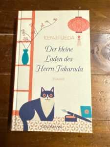 Dunkelrot Rezi Der kleine Laden des Herrn Takarada. Das Cover zeigt eine Illustration mit Katze, Stift, Buch und eine Vase.