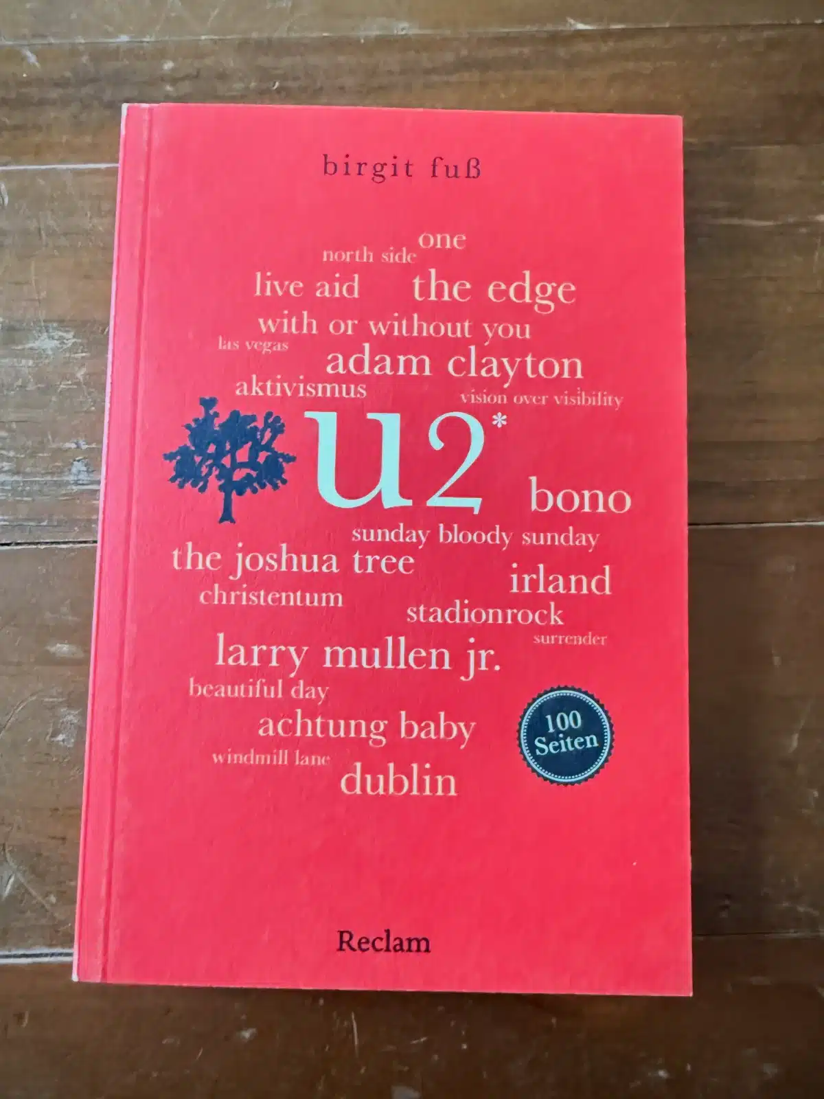U2 100 Seiten Rezension: Cover des Buches von Birgit Fuß, erschienen im Reclam Verlag, auf Holztisch fotografiert