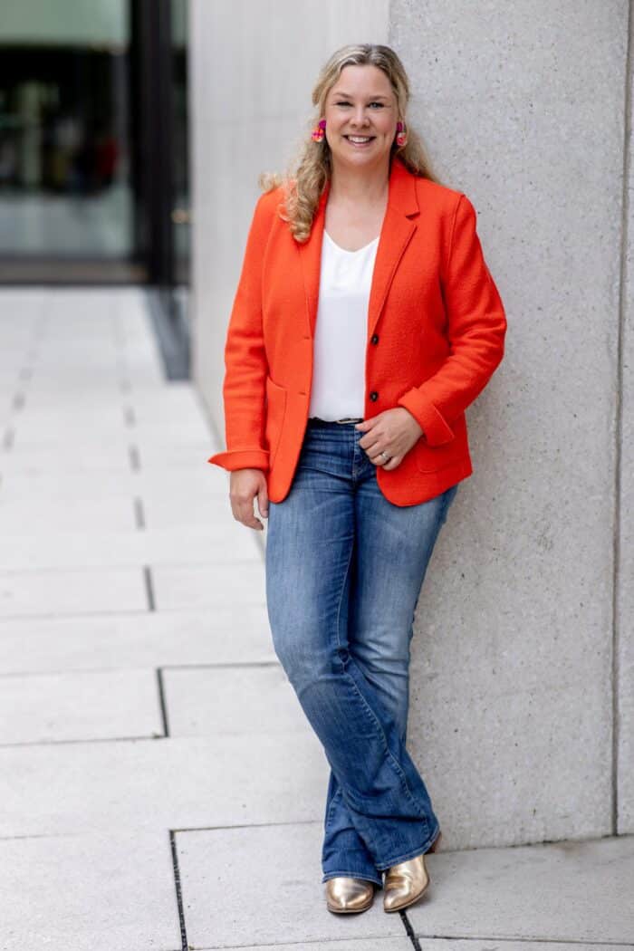 Birgit Hövener in orangefarbenem Blazer, pinken Ohrringen und goldenen Schuhen vor einer Betonwand