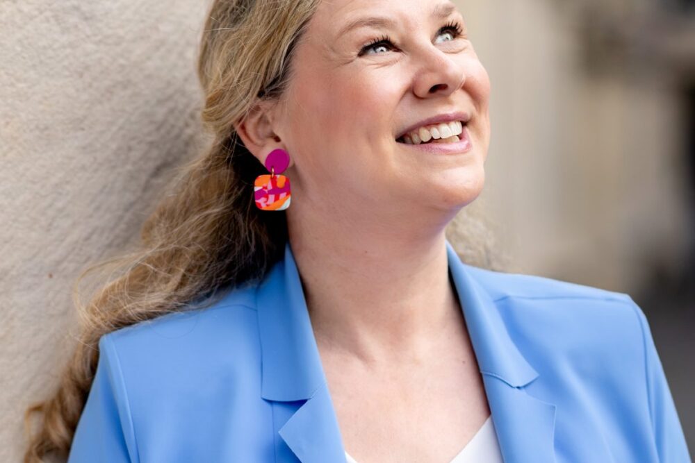 Birgit Hövener lehnt lachend an einer hellen Hauswand, hellblauer Blazer und bunte Ohrringe