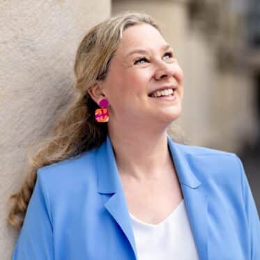 Birgit Hövener lehnt lachend an einer hellen Hauswand, hellblauer Blazer und bunte Ohrringe