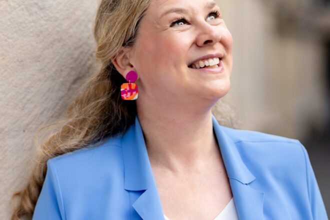 Birgit Hövener lehnt lachend an einer hellen Hauswand, hellblauer Blazer und bunte Ohrringe