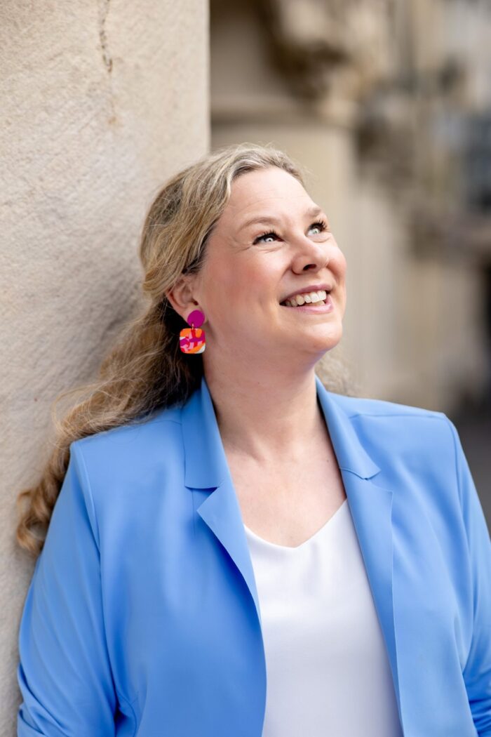 Birgit Hövener lehnt lachend an einer hellen Hauswand, hellblauer Blazer und bunte Ohrringe
